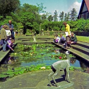 photograph of a group of schoolchildren and a couple of adults gathered around the edge of a pond. They are looking into the pond, and some of them are pointing into it. There is a stone sculpture of a doe in the foreground. 