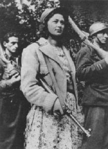 Black and white image of a woman in a dress and jacket, carrying a gun over her arm. Two men in the background also carry guns. They are all looking off camera, with calm expressions.