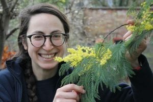 Irene Cambi, A woman with her hair in a plait and wearing glasses, smiles broadly. She is holding the foliage of a yellow feathery plant, Acacia dealbata.