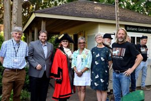 Six people are standing in a line, one has red ceremonial mayor cloak; they're standing in front of the Garden Welcome Lodge. 