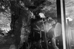 Old black and white image of a coal miner in shorts and vest drilling a seam of black coal.
