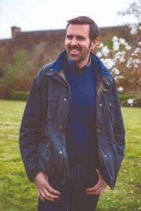 Adam Frost, dressed in navy blue stands smiling in a lawned garden.